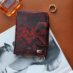 Peut inclure: Un portefeuille à motif peau de serpent rouge et noir avec une fermeture à rabat. Le portefeuille est posé sur un magazine avec un flacon de parfum et une bague dorée en arrière-plan. Le portefeuille porte le mot "DESIGN".