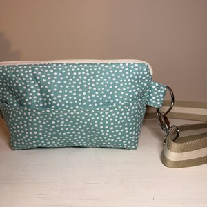 May include: A teal and white polka dot fanny pack with a cream-colored zipper and a matching adjustable strap. The strap has a beige and white striped pattern and is attached with silver metal hardware. The bag has a front pocket.