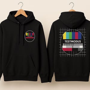 Op de afbeelding: Zwarte hoodie met een voorzak en een capuchon. De voorkant heeft een cirkelvormig logo met een kleurenstaaf en de tekst "FREE MODES". De achterkant toont een testpatroongrafiek met het woord "TESTMODUS".