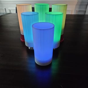 May include: Six cylindrical glass lamps with different colored lights. The lamps are arranged in a semi-circle on a dark wooden surface. The lights are blue, green, yellow, and orange.