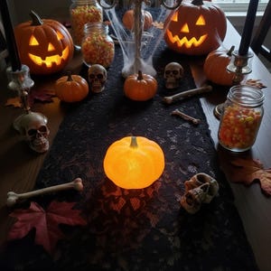 May include: A Halloween table setting with carved pumpkins, skulls, and faux bones. Lit pumpkins and candles create a spooky ambiance. Candy corn in glass jars adds a festive touch to the black lace runner.