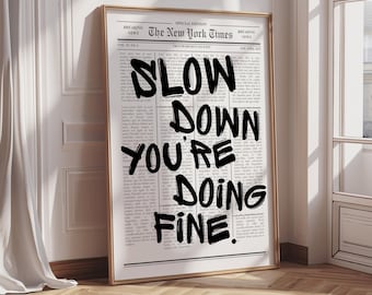 May include: A framed print featuring a vintage newspaper background with the headline "The New York Times." Bold black text overlays the newspaper, reading "SLOW DOWN YOU'RE DOING FINE."