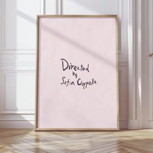 Puede incluir: Un póster enmarcado con el texto "Directed by Sofia Coppola" en escritura negra sobre un fondo rosa claro. El marco es de color marrón claro. El póster está apoyado contra una pared blanca y un suelo de madera.