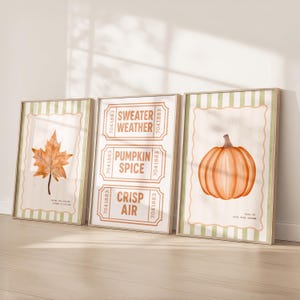 Può includere: Tre stampe artistiche incorniciate a tema autunnale. Una stampa presenta una foglia arancione, un'altra ha il testo "Sweater Weather, Pumpkin Spice, Crisp Air", e la terza mostra una zucca arancione. Tutte hanno uno sfondo beige e bordi a righe verdi.