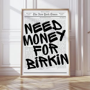 Può includere: Una stampa incorniciata con un design in stile giornale, che mostra il testo "NEED MONEY FOR BIRKIN" in grandi lettere nere. La stampa è incorniciata in una cornice marrone chiaro, con le parole "The New York Times" visibili in alto. Il design è monocromatico.