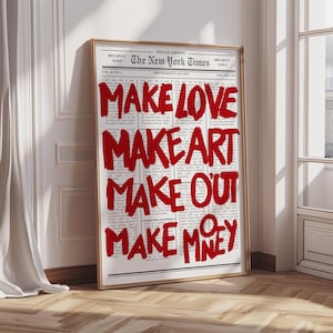 Puede incluir: Impresión enmarcada de una página del periódico New York Times con el texto rojo "MAKE LOVE MAKE ART MAKE OUT MAKE MONEY". La impresión está en un marco dorado y se apoya en una pared blanca.