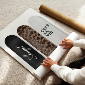 May include: A white framed art print featuring three skateboard designs. One is black with the word "Angel" in white script, another has a leopard print pattern, and the third is white with the text "C'est La Vie". A rolled-up poster is next to the art print.