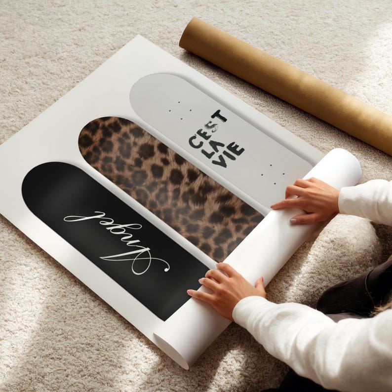 May include: A white framed art print featuring three skateboard designs. One is black with the word "Angel" in white script, another has a leopard print pattern, and the third is white with the text "C'est La Vie". A rolled-up poster is next to the art print.