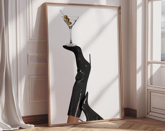 May include: Art print featuring a martini glass with olives balanced on a black high-heeled boot. The artwork is framed in gold and leans against a white wall, with a wooden floor in the foreground. The image has a minimalist, fashion-forward aesthetic.
