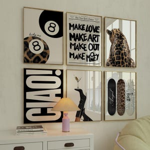 May include: A collection of framed art prints with gold frames. The prints feature various designs, including a leopard print, a martini, a pool ball, and text that reads "MAKE LOVE MAKE ART MAKE OUT MAKE MONEY", "CIAO!", "Angel", and "CEST LA VIE".