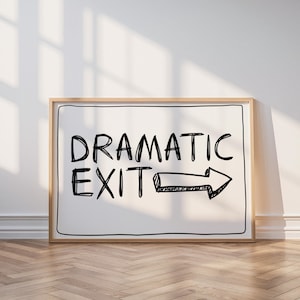 May include: A framed sign displaying the words "DRAMATIC EXIT" in black, with a hand-drawn arrow pointing right. The sign is positioned against a white wall and rests on a wooden floor.