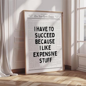 May include: A framed print resembling a newspaper front page. The headline reads "I HAVE TO SUCCEED BECAUSE I LIKE EXPENSIVE STUFF" in bold, black letters. The print is in a gold frame, leaning against a wall.