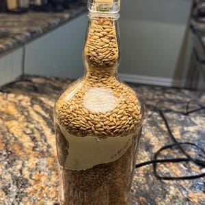Puede incluir: Una botella de vidrio transparente llena de granos, sellada con una tapa verde. La botella tiene una etiqueta con texto y est&aacute; parcialmente llena de granos marrones. La botella est&aacute; sobre una encimera.