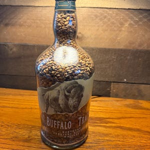 Puede incluir: Una botella de vidrio transparente llena de granos, con una tapa verde y una etiqueta con una ilustraci&oacute;n de un bisonte. La etiqueta dice "Buffalo Trace Kentucky Straight Bourbon Whiskey."