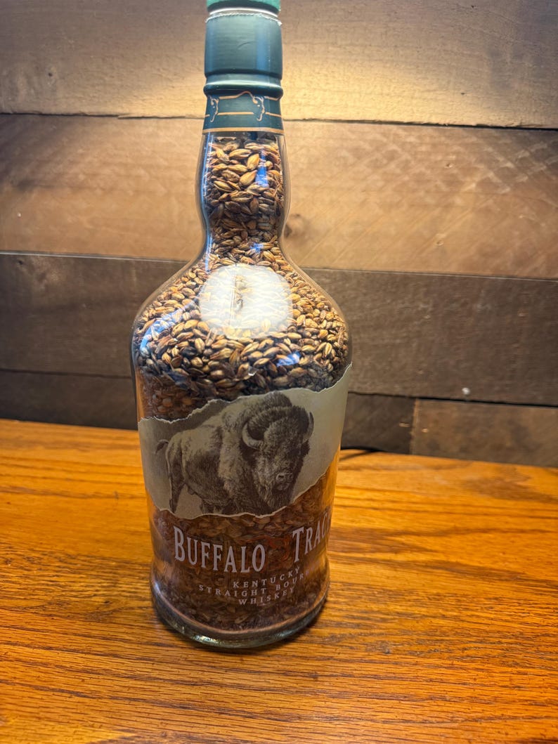 Puede incluir: Una botella de vidrio transparente llena de granos, con una tapa verde y una etiqueta con una ilustraci&oacute;n de un bisonte. La etiqueta dice "Buffalo Trace Kentucky Straight Bourbon Whiskey."
