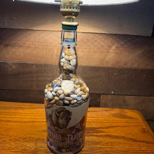 Puede incluir: Una l&aacute;mpara hecha con una botella de bourbon Buffalo Trace reutilizada, llena de peque&ntilde;as piedras. La botella tiene una etiqueta con una ilustraci&oacute;n de un bisonte. La l&aacute;mpara tiene una pantalla negra y est&aacute; sobre una superficie de madera.
