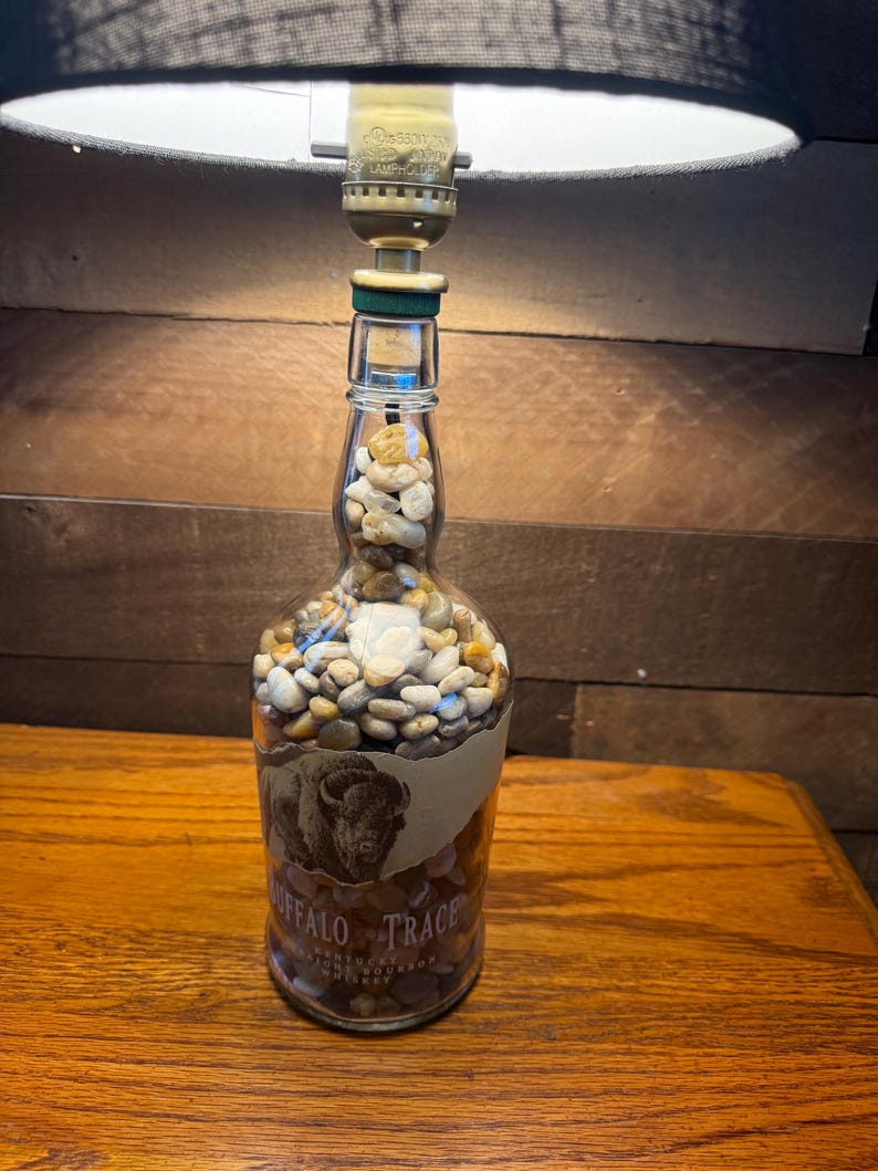 Puede incluir: Una l&aacute;mpara hecha con una botella de bourbon Buffalo Trace reutilizada, llena de peque&ntilde;as piedras. La botella tiene una etiqueta con una ilustraci&oacute;n de un bisonte. La l&aacute;mpara tiene una pantalla negra y est&aacute; sobre una superficie de madera.