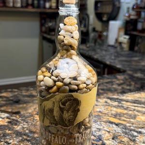 Puede incluir: Una botella de whisky Buffalo Trace de vidrio transparente llena de peque&ntilde;as piedras lisas, marrones y blancas. La botella tiene un corcho y una etiqueta con una ilustraci&oacute;n de bisonte. La botella est&aacute; sobre una encimera moteada.