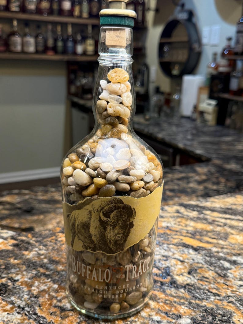 Puede incluir: Una botella de whisky Buffalo Trace de vidrio transparente llena de peque&ntilde;as piedras lisas, marrones y blancas. La botella tiene un corcho y una etiqueta con una ilustraci&oacute;n de bisonte. La botella est&aacute; sobre una encimera moteada.