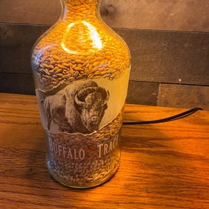 Puede incluir: Una l&aacute;mpara de botella de vidrio reutilizada llena de granos dorados. La botella presenta una etiqueta con una ilustraci&oacute;n de un bisonte y las palabras "Buffalo Trace". La l&aacute;mpara est&aacute; enchufada, emitiendo un brillo c&aacute;lido y acogedor.