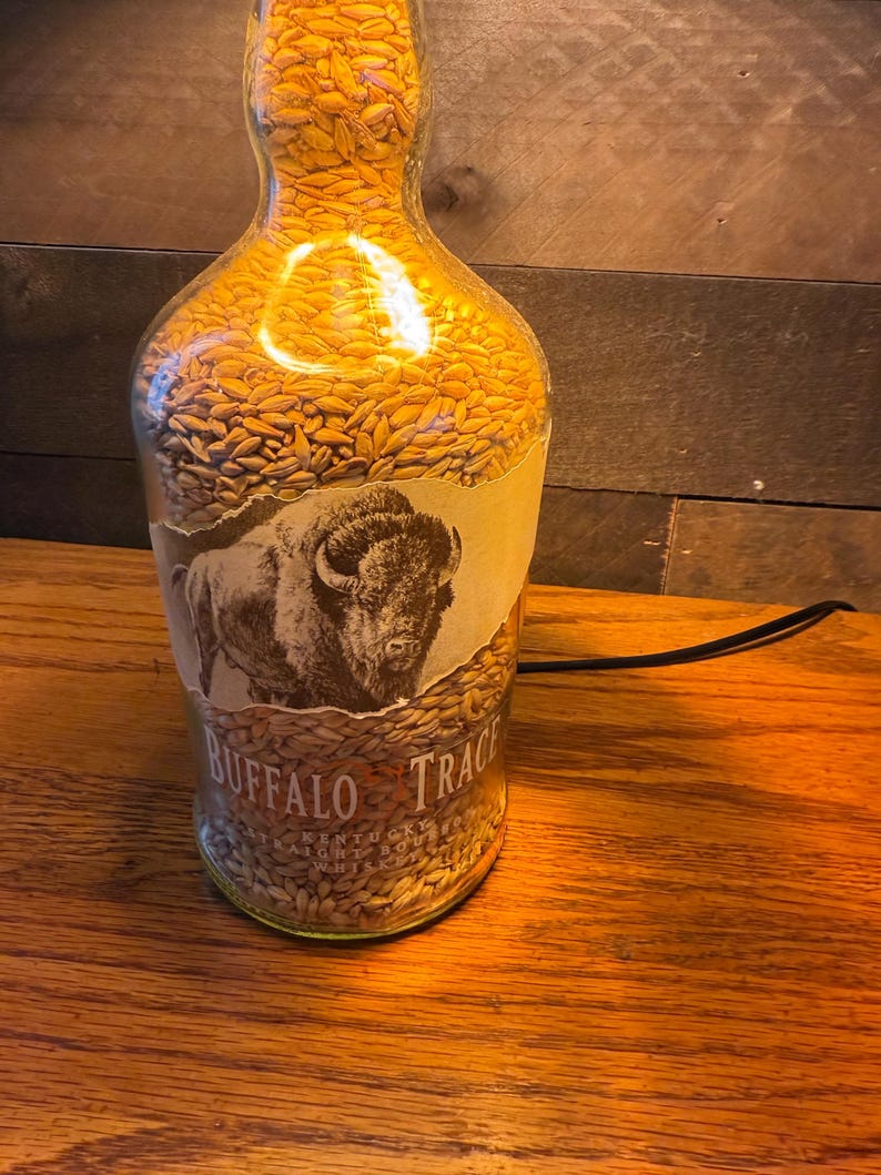 Puede incluir: Una l&aacute;mpara de botella de vidrio reutilizada llena de granos dorados. La botella presenta una etiqueta con una ilustraci&oacute;n de un bisonte y las palabras "Buffalo Trace". La l&aacute;mpara est&aacute; enchufada, emitiendo un brillo c&aacute;lido y acogedor.