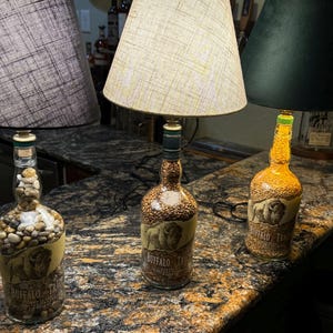 Puede incluir: Tres l&aacute;mparas decorativas hechas de botellas de licor reutilizadas. Las l&aacute;mparas presentan diferentes pantallas y rellenos de botellas, incluyendo guijarros y granos. Las botellas tienen una etiqueta con una imagen de b&uacute;falo y el texto "BUFFALO TRACE".