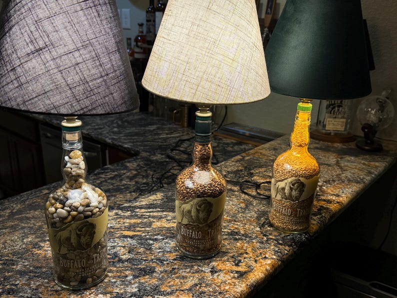 Puede incluir: Tres l&aacute;mparas decorativas hechas de botellas de licor reutilizadas. Las l&aacute;mparas presentan diferentes pantallas y rellenos de botellas, incluyendo guijarros y granos. Las botellas tienen una etiqueta con una imagen de b&uacute;falo y el texto "BUFFALO TRACE".