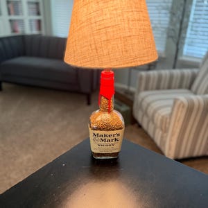 Puede incluir: Una lámpara de mesa con una pantalla de tela beige. La base es una botella de whisky Maker's Mark, sellada con cera roja. La botella está llena de granos. La lámpara está sobre una mesa negra.