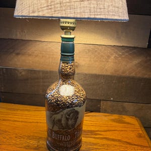 Puede incluir: Una l&aacute;mpara de mesa &uacute;nica hecha de una botella de whisky Buffalo Trace reutilizada. La botella est&aacute; llena de peque&ntilde;os granos de color marr&oacute;n claro y tiene una etiqueta con una ilustraci&oacute;n de b&uacute;falo. La l&aacute;mpara tiene una pantalla de tela beige.