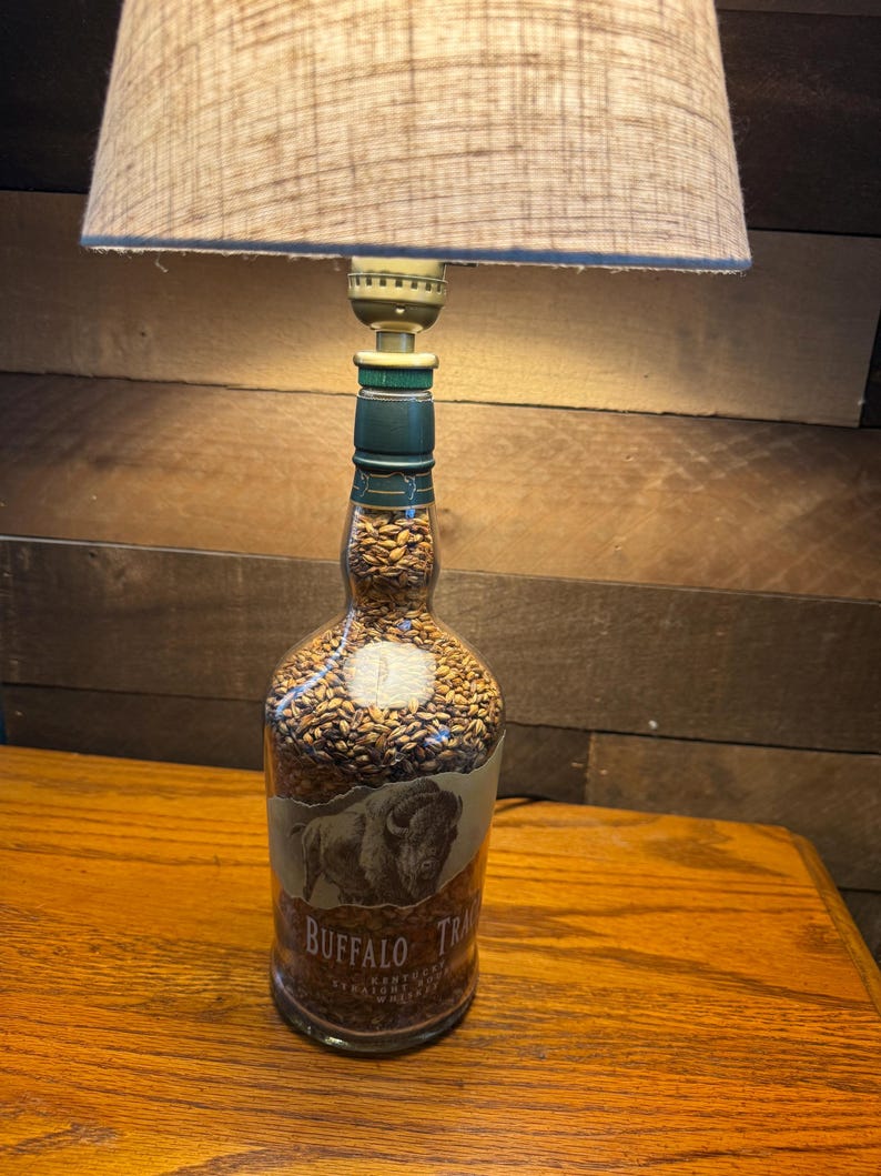 Puede incluir: Una l&aacute;mpara de mesa &uacute;nica hecha de una botella de whisky Buffalo Trace reutilizada. La botella est&aacute; llena de peque&ntilde;os granos de color marr&oacute;n claro y tiene una etiqueta con una ilustraci&oacute;n de b&uacute;falo. La l&aacute;mpara tiene una pantalla de tela beige.