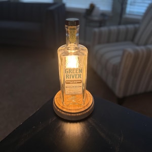 May include: A repurposed bourbon bottle lamp with the text "GREEN RIVER SINGLE BARREL BOURBON". The clear glass bottle is lit from within, resting on a wooden base. The lamp is a unique home decor item.