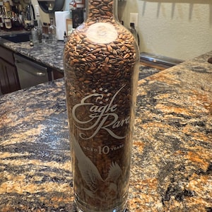 Puede incluir: Una botella de vidrio transparente llena de granos marrones, con el texto grabado "Eagle Rare Aged 10 Years" y un gráfico de águila. La botella se exhibe sobre una encimera con un patrón moteado.