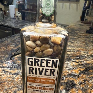 May include: A clear glass bottle filled with smooth stones, labeled "Green River Single Barrel Kentucky Straight Bourbon Whiskey." The label is brown and white with black text. The bottle is filled with stones.