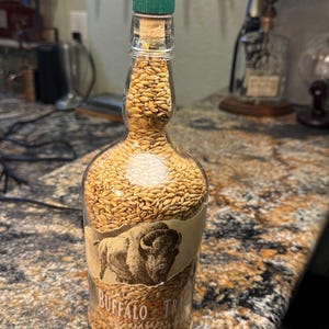 Puede incluir: Una l&aacute;mpara de botella de vidrio reutilizada llena de granos. La botella presenta una etiqueta con una ilustraci&oacute;n de un bisonte y las palabras "Buffalo Trace". La l&aacute;mpara tiene un corcho verde y un accesorio de l&aacute;mpara de lat&oacute;n.