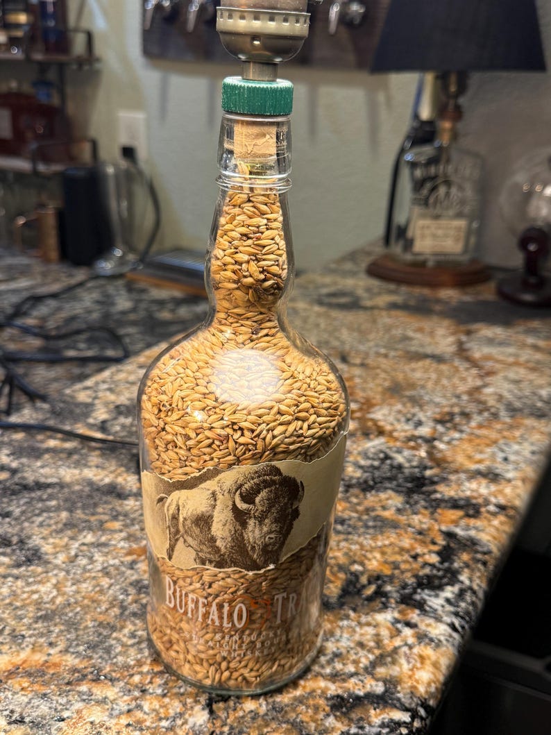 Puede incluir: Una l&aacute;mpara de botella de vidrio reutilizada llena de granos. La botella presenta una etiqueta con una ilustraci&oacute;n de un bisonte y las palabras "Buffalo Trace". La l&aacute;mpara tiene un corcho verde y un accesorio de l&aacute;mpara de lat&oacute;n.
