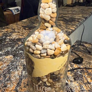 Puede incluir: Una botella de vidrio reutilizada llena de piedras de r&iacute;o lisas y multicolores. La botella tiene un tap&oacute;n de corcho y una etiqueta que dice "TRACE Bourbon". El objeto es decorativo.