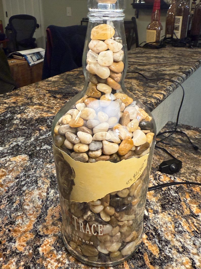 Puede incluir: Una botella de vidrio reutilizada llena de piedras de r&iacute;o lisas y multicolores. La botella tiene un tap&oacute;n de corcho y una etiqueta que dice "TRACE Bourbon". El objeto es decorativo.
