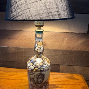 Puede incluir: Una l&aacute;mpara &uacute;nica hecha de una botella de whisky Buffalo Trace reutilizada. La botella est&aacute; llena de peque&ntilde;as piedras y presenta una etiqueta con una ilustraci&oacute;n de un bisonte. La l&aacute;mpara tiene una pantalla negra y se encuentra sobre una superficie de madera.