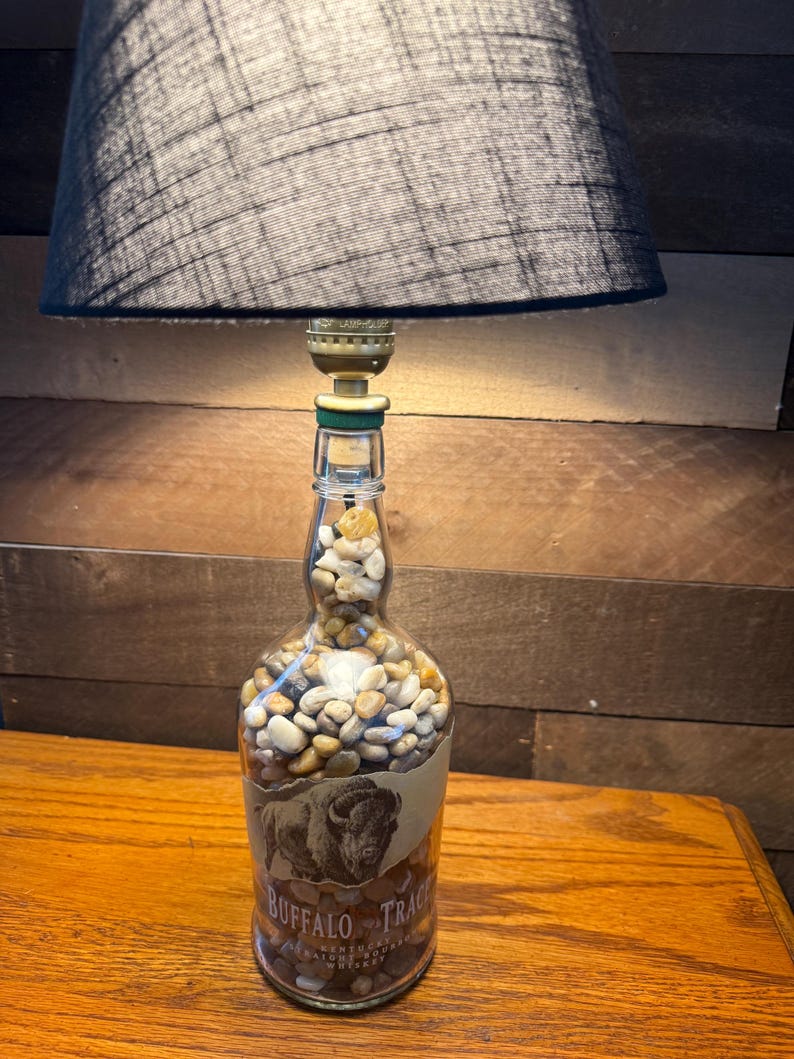 Puede incluir: Una l&aacute;mpara &uacute;nica hecha de una botella de whisky Buffalo Trace reutilizada. La botella est&aacute; llena de peque&ntilde;as piedras y presenta una etiqueta con una ilustraci&oacute;n de un bisonte. La l&aacute;mpara tiene una pantalla negra y se encuentra sobre una superficie de madera.