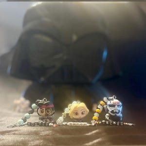 Pode incluir: Três pulseiras de contas coloridas com pingentes de personagens do universo Star Wars. Os pingentes são de Hunter, Omega e Wrecker. As pulseiras estão sobre uma superfície marrom em frente a uma imagem desfocada de Darth Vader.