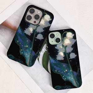 Könnte beinhalten: Zwei schwarze Handyhüllen mit floralem Design. Die Hüllen haben einen dunklen Hintergrund mit einer detaillierten Illustration von weißen Maiglöckchenblüten, grünen Blättern und funkelnden Akzenten.