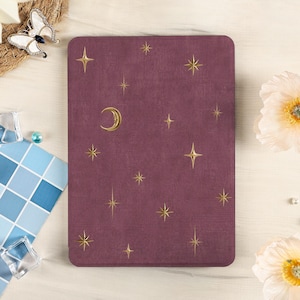 Könnte beinhalten: Eine rechteckige, weinrote Tablethülle mit einem Himmelsdesign. Goldfarbene Sterne und ein Mond sind über die Oberfläche verstreut. Die Hülle ist von dekorativen Elementen umgeben, darunter ein Schmetterling, Blumen und kleine Glaskuben.