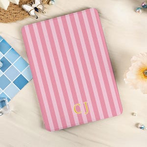 Peut inclure: Une housse de liseuse électronique rectangulaire présentant un motif à rayures verticales dans des tons alternés de rose clair et foncé. Le bas du centre de la housse affiche les lettres capitales CJ en doré avec une police serif classique.