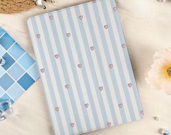 Puede incluir: Una funda rectangular para lector electrónico con un patrón de rayas verticales en azul claro y blanco, decorada con pequeños corazones rosas dibujados a mano dispersos por la superficie. La funda tiene un acabado liso y esquinas redondeadas.