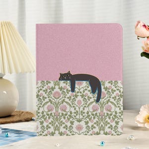 May include: A pink notebook featuring a black cat illustration. The top section is plain pink, and the bottom has a floral pattern with pink flowers and green leaves. The cat is depicted in a relaxed pose.