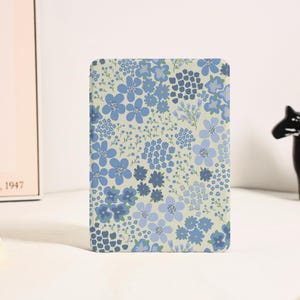 Peut inclure: Une housse de liseuse rectangulaire ornée d'un motif répétitif de petites fleurs dans des tons bleu clair, bleu moyen et bleu marine foncé sur un fond crème pâle. Le motif floral recouvre toute la surface avant de l'objet.