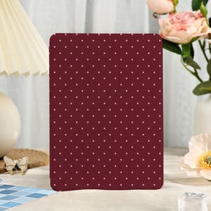 May include: A rectangular tablet or e-reader case featuring a deep burgundy background with a repeating pattern of small, light pink polka dots. The case has rounded corners and a smooth, matte finish, displaying a consistent geometric design.