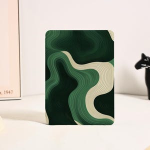May include: A rectangular card with a layered, abstract design in shades of green and beige. The artwork features curved lines creating a topographic effect. The card is set against a white background, with a black horse figurine visible.