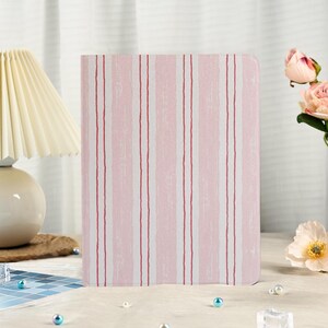 Könnte beinhalten: Ein Notizbuch mit rosa und weiß gestreiftem Einband. Das Design zeigt vertikale Streifen in Rosa, Weiß und dünnen roten Linien. Das Notizbuch steht aufrecht. Im Hintergrund eine Lampe und Blumen.