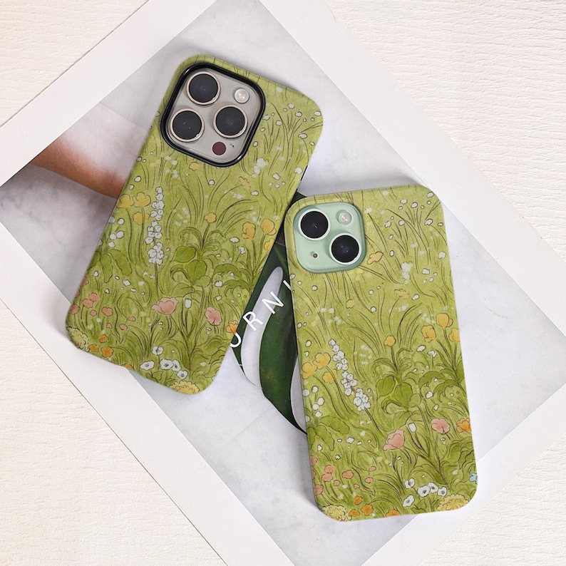 May include: Two phone cases with a green floral design. The cases feature a pattern of wildflowers in shades of yellow, pink, and white against a green background. The cases have camera cutouts and are displayed on a white surface.
